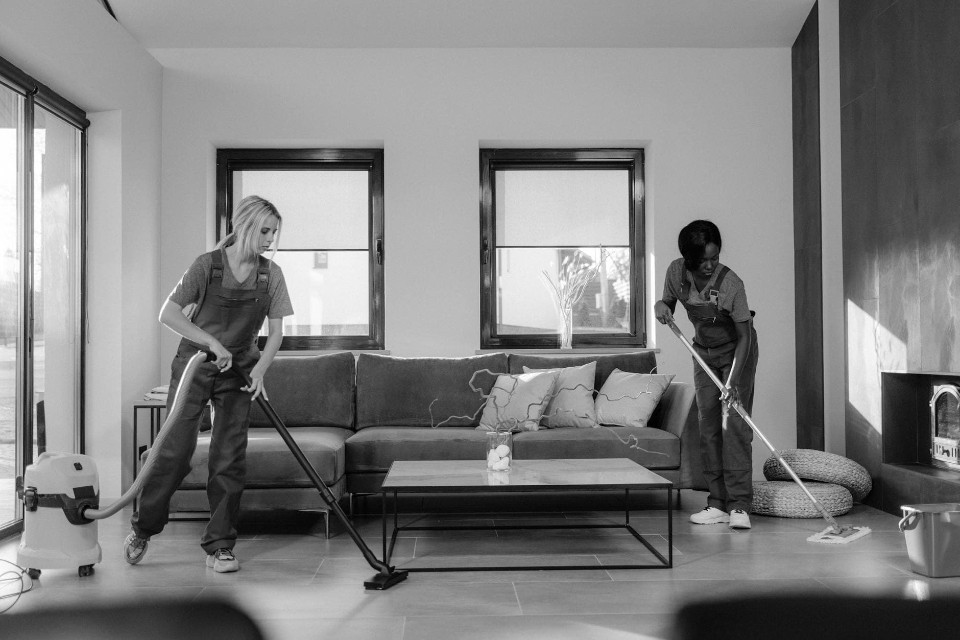 Two Long Island Maid cleaners in work uniforms using a vacuum and mop in a modern Long Island living room — Office Cleaning in Port Washington, Nassau County